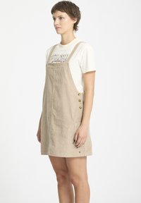 Beige corduroy jurk met een licht uitlopende vorm, voorzien van verstelbare bandjes, zijknopen en voorzakken. Draag het over een wit T-shirt.