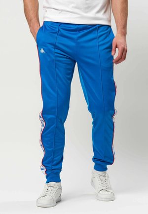 Male wearing blue athletic pants with white and red side stripes and white sneakers, standing with hands relaxed by sides.