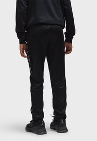 Person in schwarzen Sporthosen mit reflektierenden Chevron-Streifen an den Seiten, schwarzen Turnschuhen mit reflektierenden Details, stehend vor weißem Hintergrund.