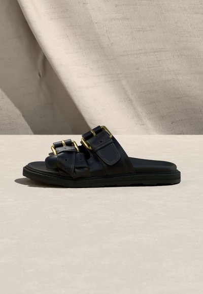 Μαύρο σανδάλι slide με δύο φαρδιές λωρίδες που διαθέτουν μεγάλα χρυσά αγκράφες, τοποθετημένο σε ανοιχτό μπεζ επιφάνεια με υφασμάτινο φόντο.