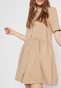 Abito beige a maniche corte con chiusura a bottoni, vita arricciata e gonna svasata. Realizzato in tessuto leggero con una trama liscia.