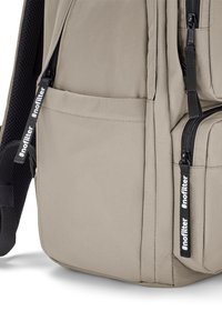Beige Rucksack mit strukturiertem Stoff, mehreren Reißverschlussfächern und einer seitlichen Netztasche mit einem schwarzen "#nofilter"-Label.