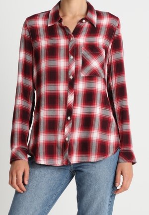 Vrouw draagt een geruit overhemd in rood, zwart en wit met borstzak, gecombineerd met een blauwe spijkerbroek, staand tegen een effen achtergrond.