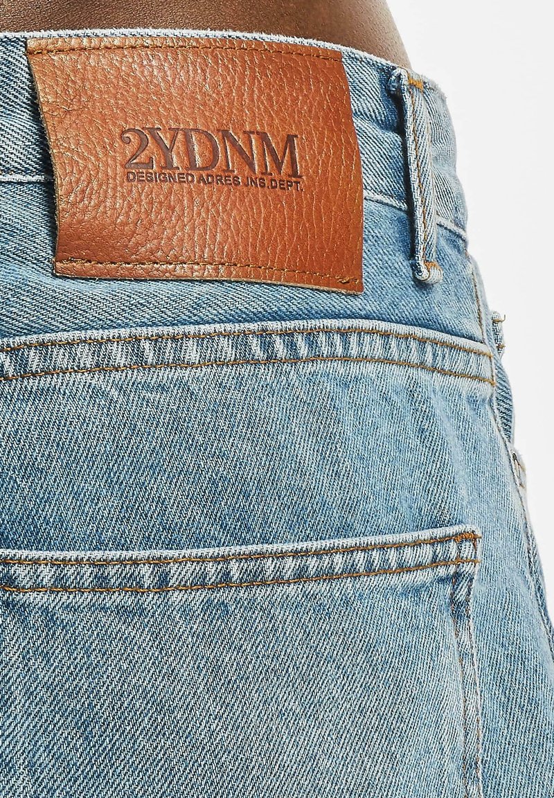 Jeans aus Denim mit einer hellblauen Waschung, versehen mit einem braunen Lederpatch mit geprägtem Text auf der Rückseite, sichtbaren Nähten und standardmäßigen Taschen.