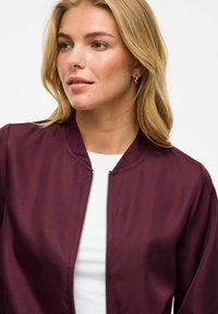 Burgunderfarget bomberjakke med en jevn tekstur, stående krage og frontglidelås. Båret over en hvit topp med subtile sømdetaljer.