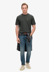 T-shirt verde scuro a maniche corte con taschino sul petto, abbinata a jeans blu chiaro. Una camicia a quadri nera e grigia legata in vita e sneakers bianche.
