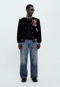 Zwarte varsity-jas met witte strepen, groot "F"-patch, gecombineerd met losse blauwe spijkerbroeken en zwarte laarzen. Geaccessoreerd met donkere zonnebrillen.