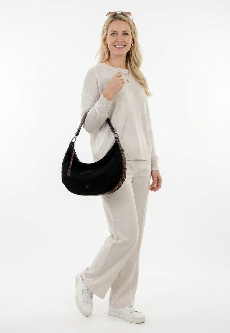 Šviesiaplaukė moteris dėvi smėlio spalvos megztinį ir kelnes, baltus sportinius batelius, šypsosi, laiko juodą rankinę su leopardiniu raštu ant balto fono.