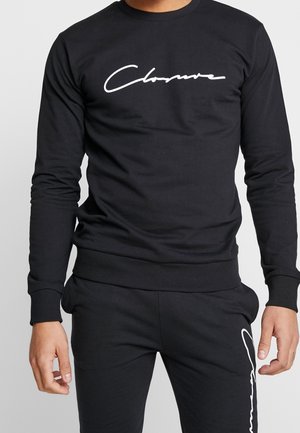 Homme portant une chemise noire à manches longues et un pantalon avec un logo cursif blanc "Closure" sur la poitrine de la chemise et la jambe du pantalon.