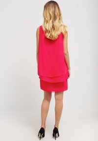 Sukienka bez rękawów w intensywnym różowym kolorze z warstwowym wzorem; przezroczysty tył, gładka tekstura i długość do kolan, w połączeniu z czarnymi butami na wysokim obcasie.
