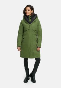 Groene winterjas met een grote zwarte capuchon, voorzien van knopen en zakken, gemaakt van een gladde stof. G gedragen met zwarte jeans en laarzen.