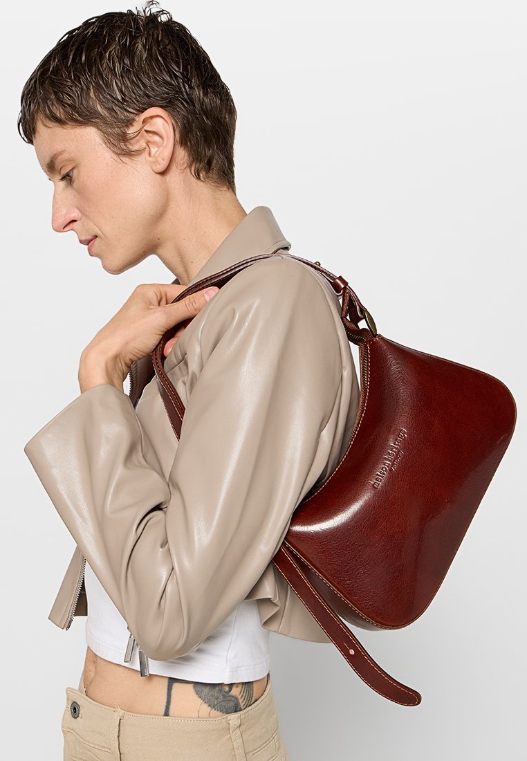 Braune Lederhandtasche mit geschwungener Form und einem einzelnen Schultergurt, kombiniert mit einer beigen Kunstlederjacke.