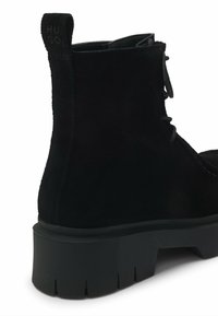Botină din suede negru, cu talpă groasă cu crampoane, șiret la față și tab de mână cu brand la toc.