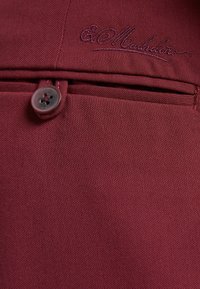 Il tessuto rosso scuro presenta una superficie liscia con un pulsante visibile e dettagli cuciti. Il nome del marchio ricamato appare sopra la tasca.