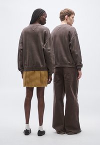 Sudaderas marrones extragrandes en dos personas, una lleva una falda amarilla a cuadros y la otra pantalones marrones de pierna ancha. Textura de tela suave.