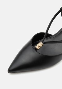 Chaussure en cuir noire à bout pointu avec une fine bride et un petit charm doré décoratif au centre avant.