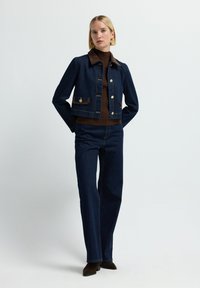 Chaqueta de mezclilla en azul oscuro con cuello y acentos de bolsillo marrón, con botones en tono dorado. Combinada con jeans de pierna ancha azul oscuro.
