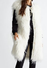 Langer weißer Kunstfell-Westen mit einer wuscheligen Textur, ausgestattet mit einem sauberen Revers und Knöpfen, getragen über einem schwarzen Outfit.