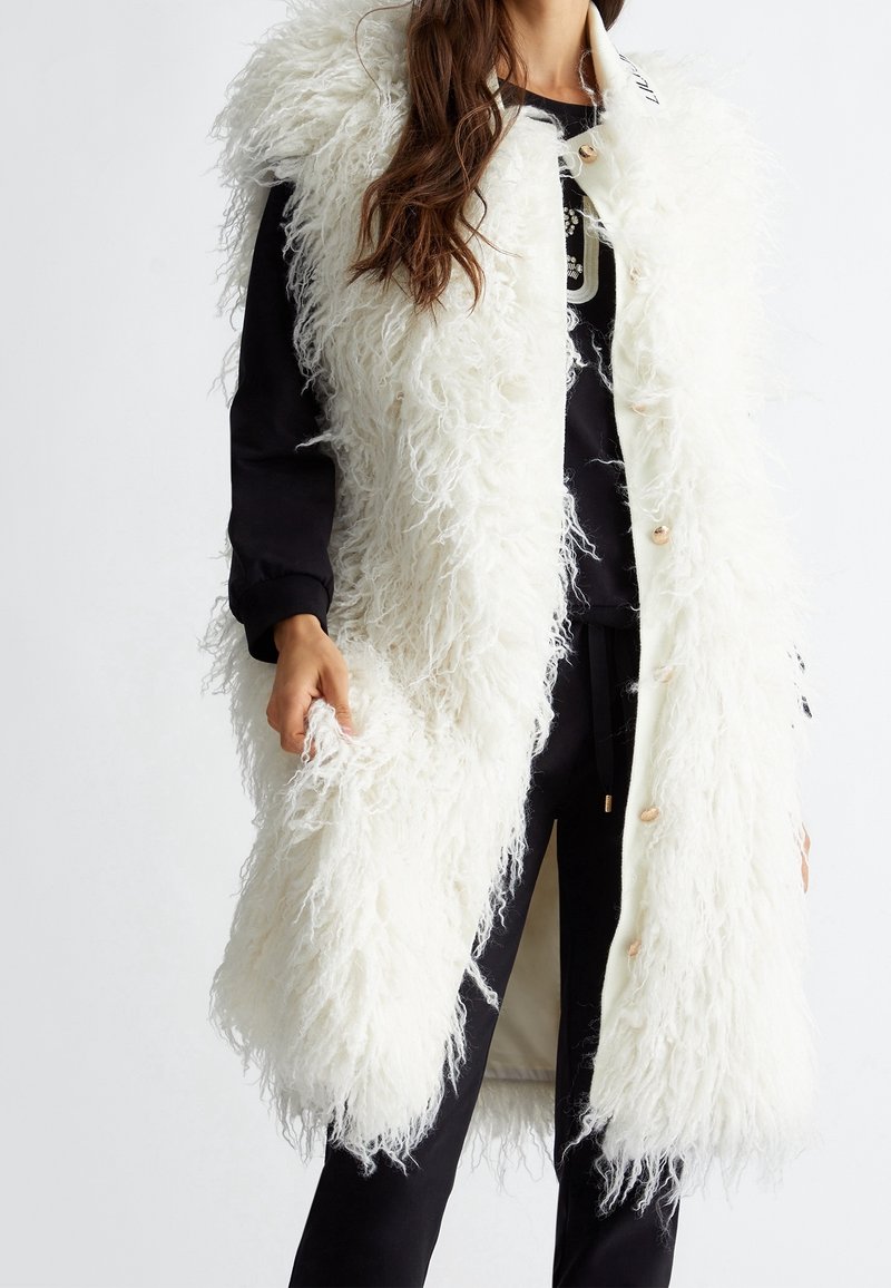 Langer weißer Kunstfell-Westen mit einer wuscheligen Textur, ausgestattet mit einem sauberen Revers und Knöpfen, getragen über einem schwarzen Outfit.