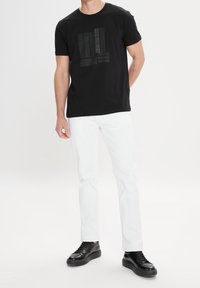 Sort T-shirt med korte ærmer og tekstureret grafisk design på forsiden, parret med hvide lige bukser og sorte sneakers.