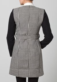 Valkoinen ja musta houndstooth-kuvioinen takki, jossa on pitkät mustat hihat ja vyötärövyö; siinä on sileä rakenne ja muotoiltu leikkaus.