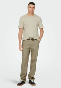 Beige Crewneck-T-Shirt, beige Chinos, brauner Gürtel, schwarze Sneakers mit weißen Schnürsenkeln, vor einem schlichten hellen Hintergrund.