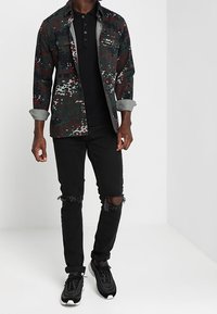 Kamouflage-mönstrad skjortjacka i grön, röd och grå; svart polo under; slitna svarta jeans; svarta sneakers med vita sulor.