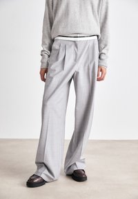Helle graue, plissierte Hosen mit einer taillierten Passform, aus einem strukturierten Stoff und mit einem anschmiegsamen Bund. Kombiniert mit dunklen, glänzenden Schuhen.