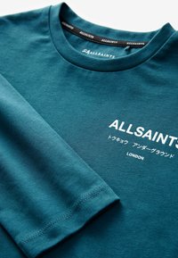 Tirkizna majica dugih rukava od pamuka. Ima okrugli izrez i bijeli tiskani natpis: "ALLSAINTS" i "LONDON", s glatkom teksturom.