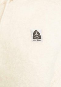 Tela de forro color crema con una textura suave, que presenta una etiqueta cuadrada con un diseño de hoja y el texto "JUST RHYSE" en el centro.