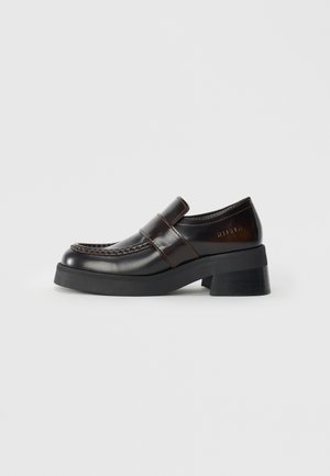 Mocasines de cuero marrón con un tacón negro grueso, que presentan un diseño cosido y una correa en la parte superior para un detalle adicional.