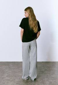 Femme aux longs cheveux blonds se tient de dos, portant un t-shirt noir à manches courtes et un pantalon gris rayé ample sur un sol gris.
