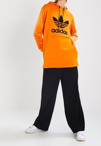 Person som bär en ljusorange Adidas-hoodie, lösa svarta byxor och vita sneakers, står med händerna i fickorna mot en enkel bakgrund.