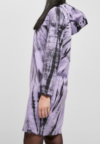 Fioletowa sukienka z kapturem w tie-dye z długimi rękawami, luźnym krojem i czarnymi akcentami tworzącymi kontrastowy wzór. Miękka faktura materiału.