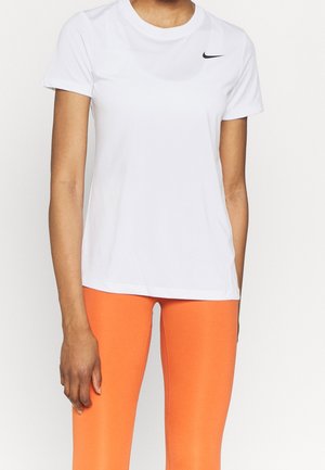 Persoon draagt een wit Nike T-shirt met korte mouwen en feloranje legging, staat met de linkerarm ontspannen langs het lichaam.