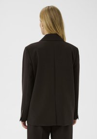 Donna bionda di spalle che indossa un blazer oversize marrone scuro con le maniche abbottonate su uno sfondo bianco semplice.