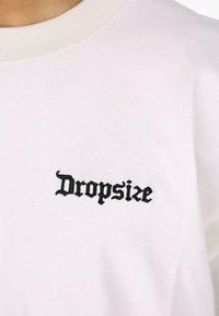 Hvit bomulls t-skjorte med et svart brodert "Dropsize"-logo på venstre brystområde. Rund hals med ribbestrikket finish.