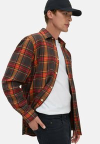 Geruite flanellen shirt in donkergrijs, oranje, rood en geel. Lange mouwen, knoopsluiting aan de voorkant, gedragen over een wit T-shirt, met een donkere broek.