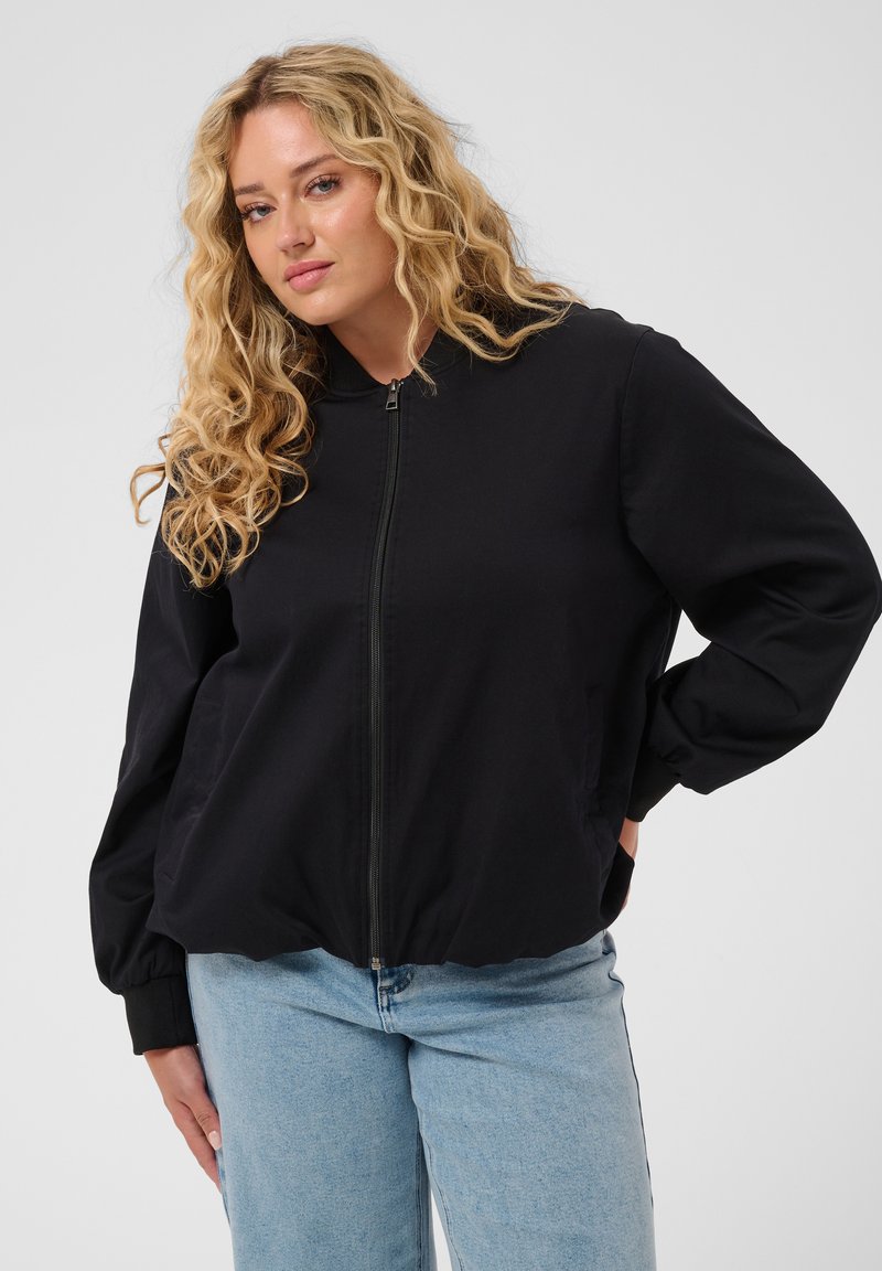 Femme aux longs cheveux blonds bouclés portant une veste noire zippée et un jean bleu clair sur un fond gris uni.