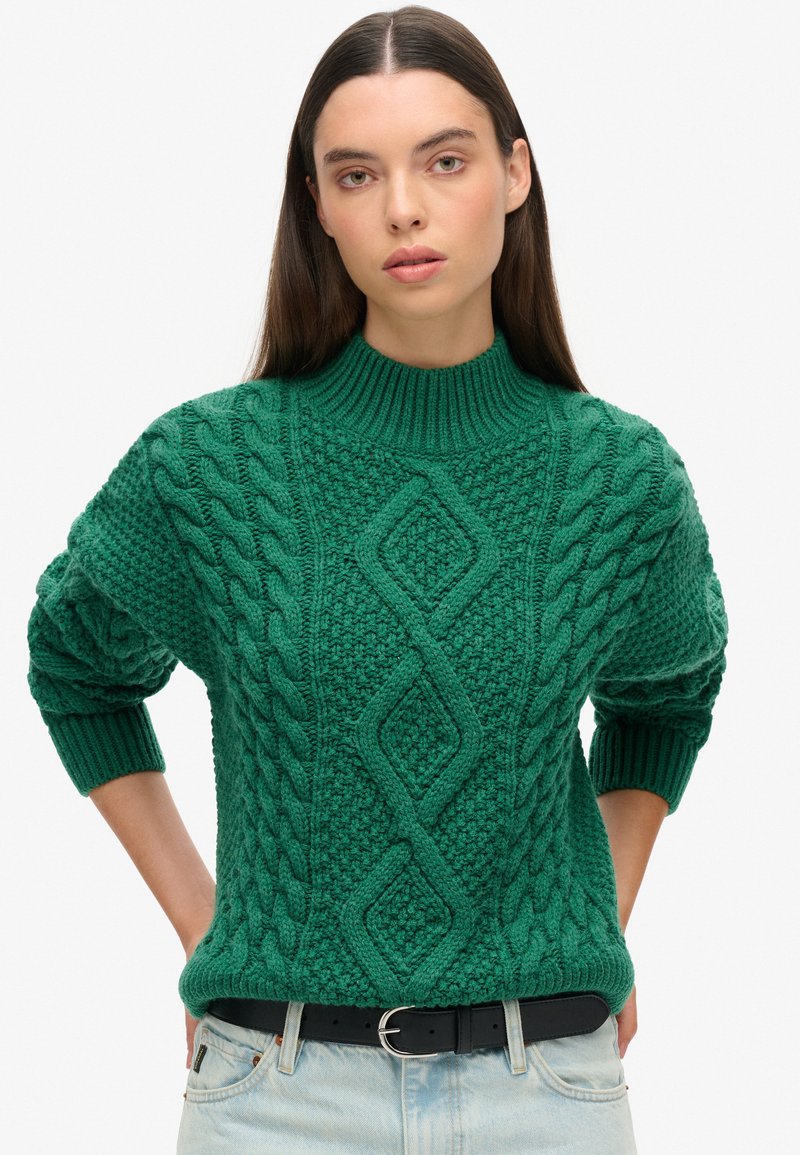 Pull en tricot vert avec un col haut, présentant des motifs en câble et un design en losange. Poignets et ourlet côtelés, assorti à une ceinture noire.