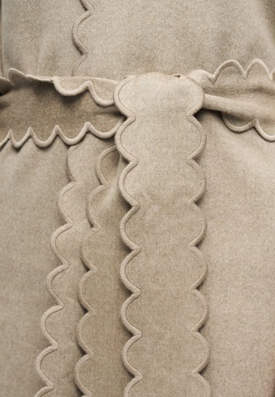 Μπεζ ύφασμα με κυματιστά άκρα και ανάγλυφες ραμμένες λεπτομέρειες που δημιουργούν ένα υφασμένο, κυματιστό μοτίβο.