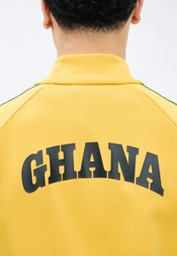 Άτομο που φαίνεται από πίσω φοράει ένα κίτρινο μπουφάν με ψηλό γιακά και τη λέξη "GHANA" τυπωμένη με μεγάλα μαύρα γράμματα στην πλάτη.