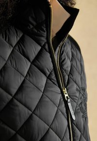 Čierna prešívaná bunda s vysokým golierom, vybavená zlatým zipsom a jemným vyšívaným zdobením. Hladká textúra a priliehavý strih.