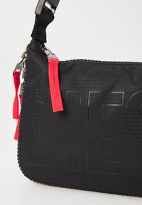 Black fabric shoulder bag with a textured surface, featuring large embossed text, red straps, and silver hardware accents.
