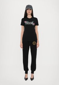 Juodas marškinėliai su gėlių "Versace Jeans Couture" logotipu, derinami su juodomis sportinėmis kelnėmis, turinčiomis auksinį apskritą logotipą. Į komplektą įeina kepuraitė ir kulniniai batai.