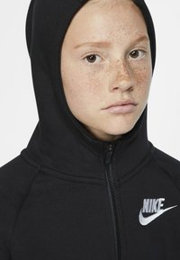 Črna jakna s kapuco na zadrgo in visokim ovratnikom, z belim logotipom Nike na levem prsnem delu. Gladka tkanina in prilegajoča se zasnova.