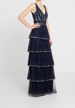 Femme portant une robe longue bleu marine sans manches à volants, avec un corsage orné de sequins et un ourlet festonné, debout, main sur la taille.