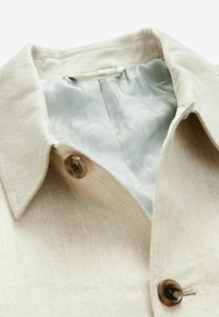 Beige stoffen jack met een puntige kraag, twee bruine knopen en een gladde lichtgrijze gestreepte voering in het kraaggebied.