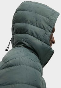 Waterdichte pufferjas in donkergroen met een gewatteerd design. Beschikt over een volledige capuchon, zijrits en druppels op de stof.