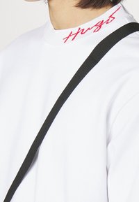 Bílá mikina s vysokým límcem, na které je červený vyšívaný text. Částečně je viditelný černý ramenní popruh, který dodává kontrastní detail.